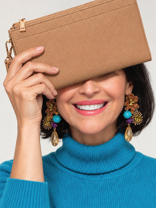 A smiling woman in a bright blue turtleneck sweater holds a tan ppurse in front of her eyes. She is wearing large, colorful statement earrings with turquoise, gold, and brown details, set against a plain light gray background.