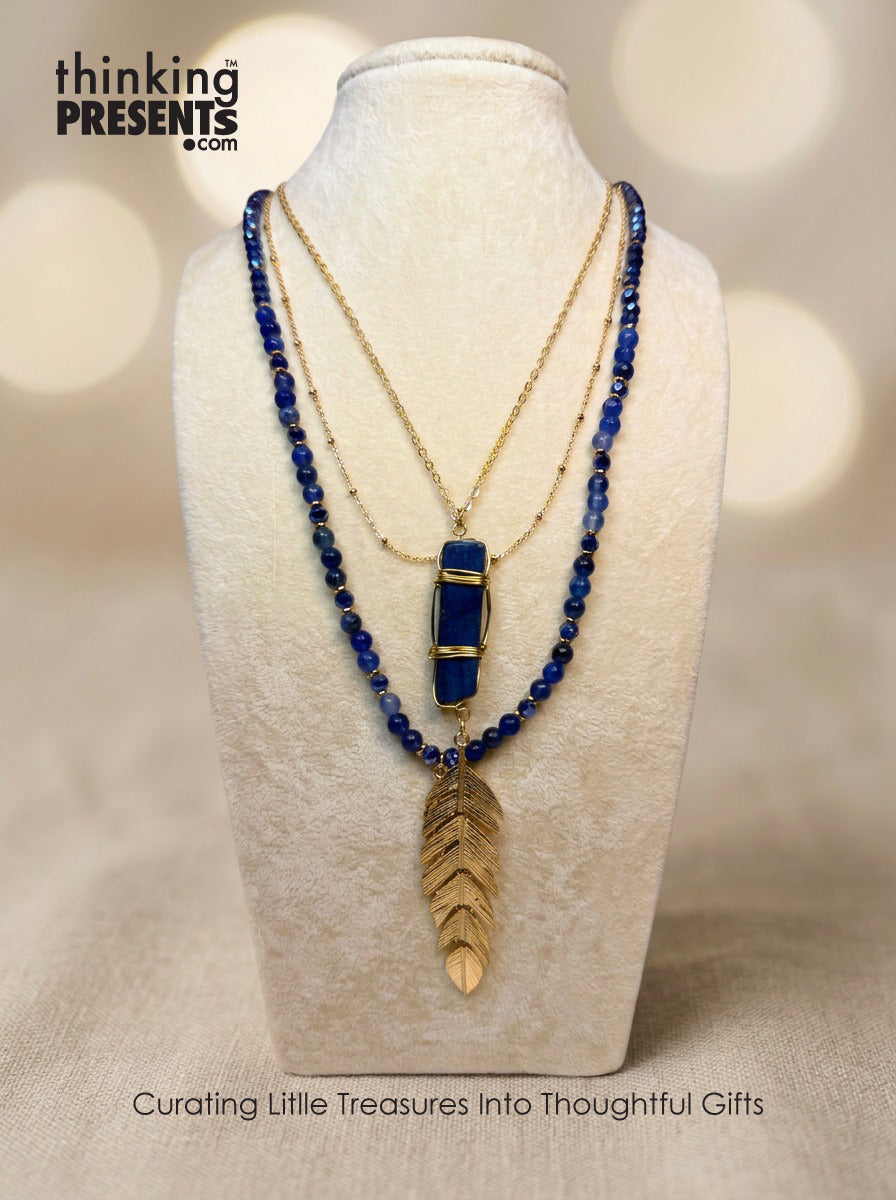 Layered gold-tone necklace with deep blue stone pendant, beaded blue strand, and feather charm, styled on a neutral display, elegant and boho-inspired curated gift aesthetic