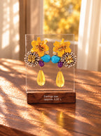 A pair of colorful statement earrings displayed on a clear acrylic stand with a wooden base. The earrings feature bright yellow flower-shaped elements, turquoise blue beads, purple beads, and gold-tone metal details, with teardrop yellow drops.