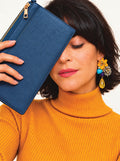 A woman in a mustard yellow ribbed turtleneck sweater holds a blue pebbled leather clutch purse next to her face. She is wearing a colorful statement earring with yellow, blue, and gold elements.