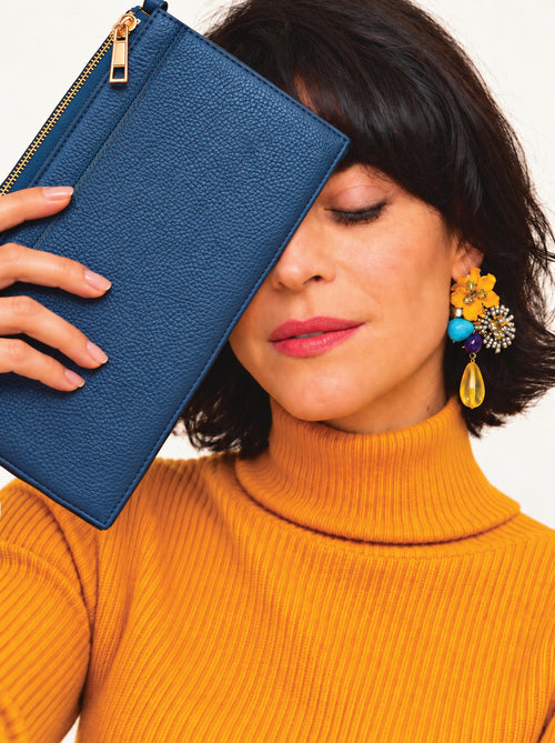 A woman in a mustard yellow ribbed turtleneck sweater holds a blue pebbled leather clutch purse next to her face. She is wearing a colorful statement earring with yellow, blue, and gold elements.