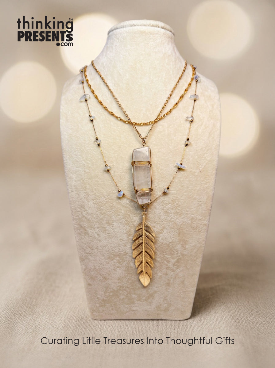 Layered gold-tone necklace with clear stone pendant, delicate beaded accents, and feather charm, styled on a neutral display, boho-inspired and feminine curated gift aesthetic