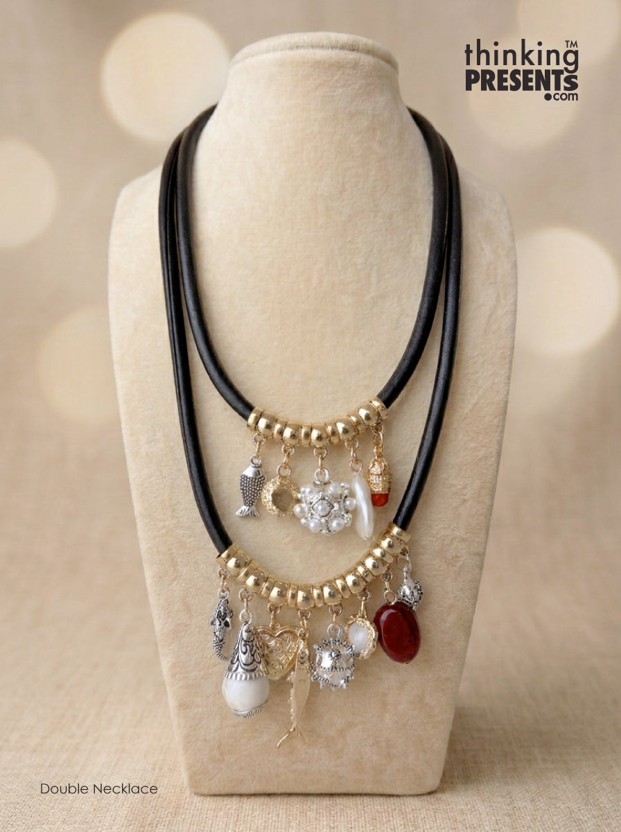 A double-strand necklace displayed on a beige fabric jewelry bust. The necklace consists of black cords with gold-tone bead accents and dangling charms in silver, white, and red.