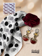 A styled flat-lay featuring a white scarf with a black and gray polka dot and brushstroke pattern, a deep burgundy velvet scrunchie, and a pair of dangling earrings with cream floral tops, amber oval stones, and red teardrop charms. A small inset shows the scarf design with text reading 'Scarf Design 25 in x 25 in.