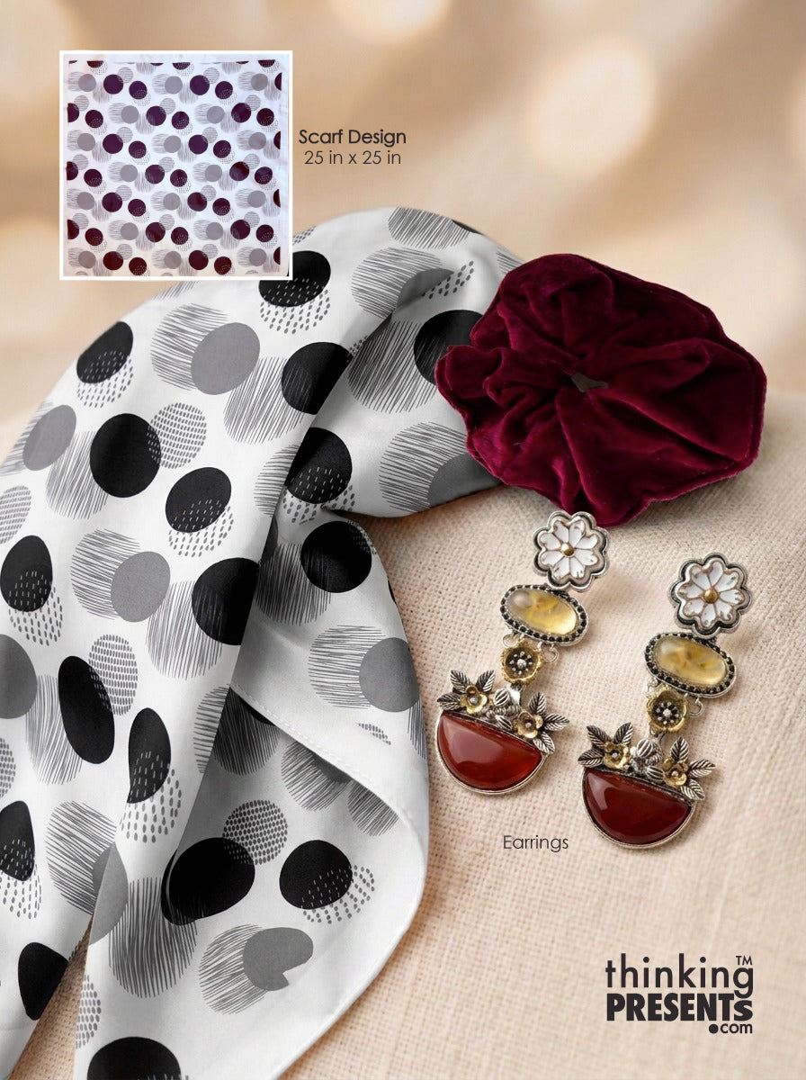 A styled flat-lay featuring a white scarf with a black and gray polka dot and brushstroke pattern, a deep burgundy velvet scrunchie, and a pair of dangling earrings with cream floral tops, amber oval stones, and red teardrop charms. A small inset shows the scarf design with text reading 'Scarf Design 25 in x 25 in.