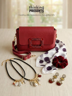 A red textured leather crossbody handbag with a studded rectangular clasp, displayed on a light beige surface with a matching red scrunchie, a white scarf with dark purple polka dots, and gold-tone jewelry. The text 'thinking PRESENTS .