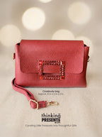 A red pebbled leather crossbody bag with a rectangular buckle embellished with rhinestones, shown on a neutral background with bokeh lights. Text overlays describe the bag and display the brand 'thinking PRESENTS'.