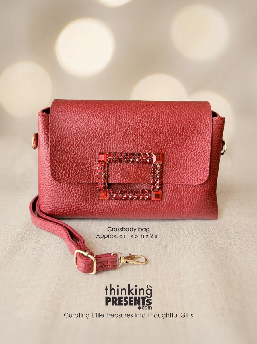A red pebbled leather crossbody bag with a rectangular buckle embellished with rhinestones, shown on a neutral background with bokeh lights. Text overlays describe the bag and display the brand 'thinking PRESENTS'.