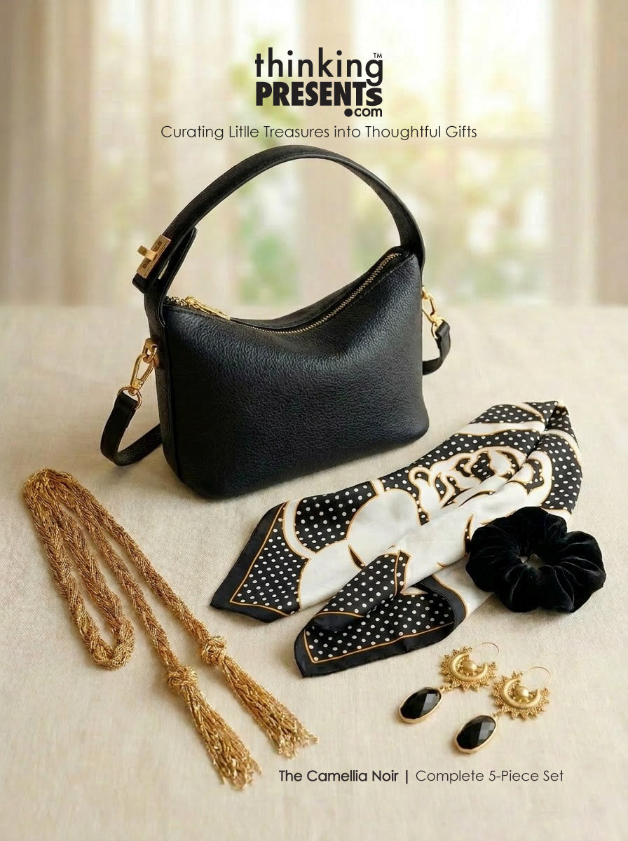 A black pebbled leather handbag displayed with a matching black, white, and gold 5-piece gift set on a light beige fabric surface. The set includes a patterned scarf, a black velvet flower clip, a gold chain with tassels, and gold earrings.