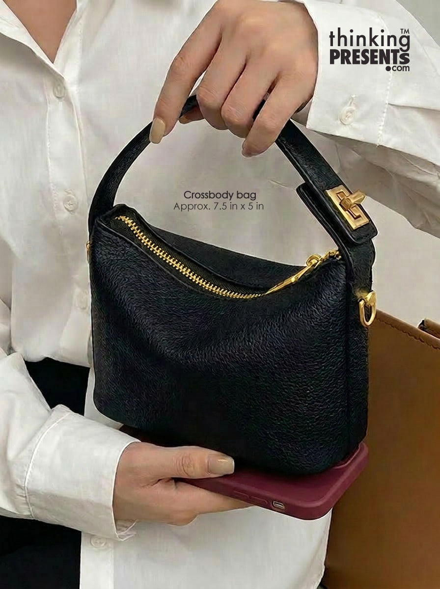 A person in a white button-up shirt holds a small black pebbled leather crossbody bag with gold-tone hardware and a top handle. The bag has a zippered top and is shown against a white shirt background with a brown surface at the lower right.