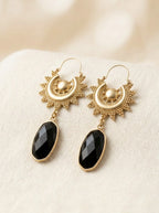 A pair of gold-toned dangle earrings with sunburst and crescent moon motifs, featuring faceted black teardrop-shaped stones, displayed on a textured cream fabric background.