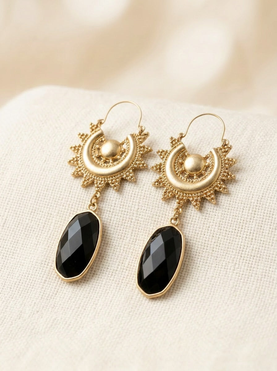 A pair of gold-toned dangle earrings with sunburst and crescent moon motifs, featuring faceted black teardrop-shaped stones, displayed on a textured cream fabric background.
