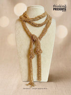 A textured gold-toned multi-strand necklace with knotted details and long tassels, displayed on a cream-colored jewelry bust against a warm beige bokeh background. The 'thinking PRESENTS' logo is visible in the top right corner.