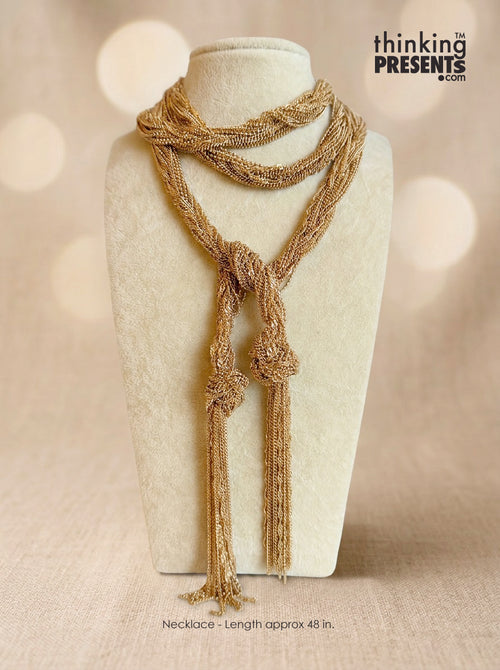 A textured gold-toned multi-strand necklace with knotted details and long tassels, displayed on a cream-colored jewelry bust against a warm beige bokeh background. The 'thinking PRESENTS' logo is visible in the top right corner.