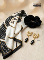 A styled flat-lay featuring a black and white silk scarf with gold detailing, a black velvet scrunchie, and a pair of gold drop earrings with black teardrop stones. The items are arranged on a beige textured fabric background with a warm bokeh backdrop.