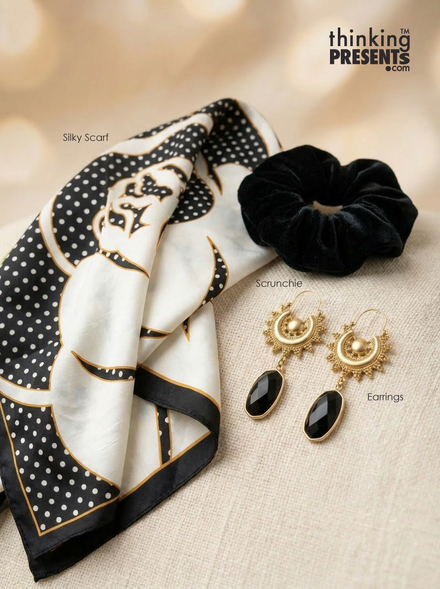 A styled flat-lay featuring a black and white silk scarf with gold detailing, a black velvet scrunchie, and a pair of gold drop earrings with black teardrop stones. The items are arranged on a beige textured fabric background with a warm bokeh backdrop.