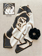 A folded scarf with a cream and black floral design, black polka dot patterned corners, and gold trim, displayed on a beige textured fabric background next to a black scrunchie. A small graphic in the top left shows the scarf design details with text reading 'Scarf Design 23 in x 23 in'.