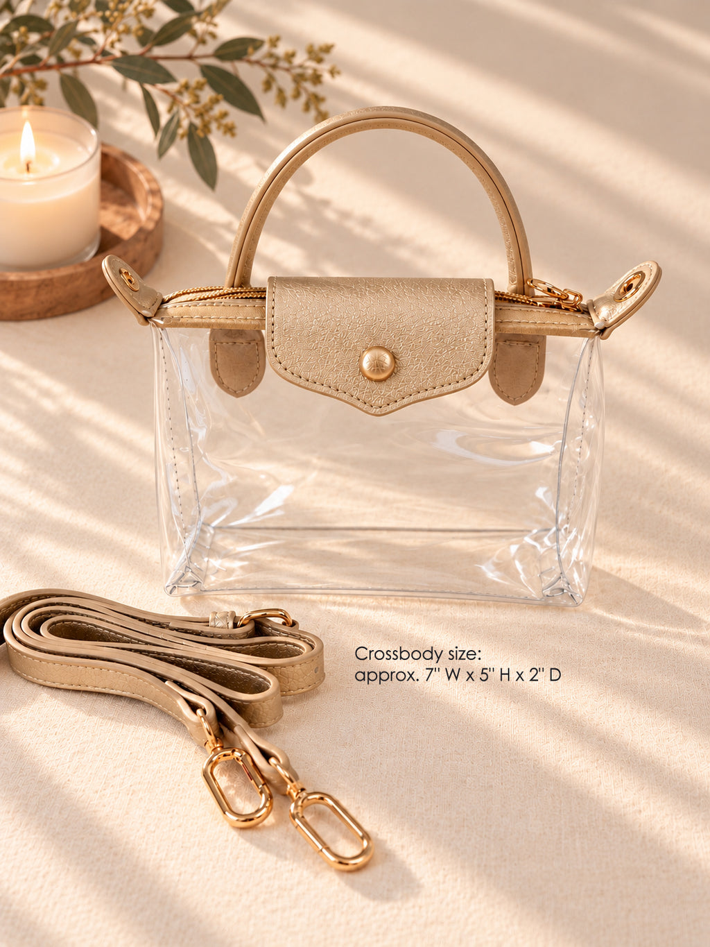 A clear acrylic crossbody handbag with a beige flap and handle, gold-tone hardware, and a matching beige detachable strap laid beside it on a warm beige fabric surface. A candle and dried leaves appear in the background