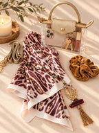A styled flat-lay featuring a sheer leopard-print scarf with a white base and dark burgundy spots, styled with a gold handbag, velvet scrunchie, phone, and candle on a beige fabric background.