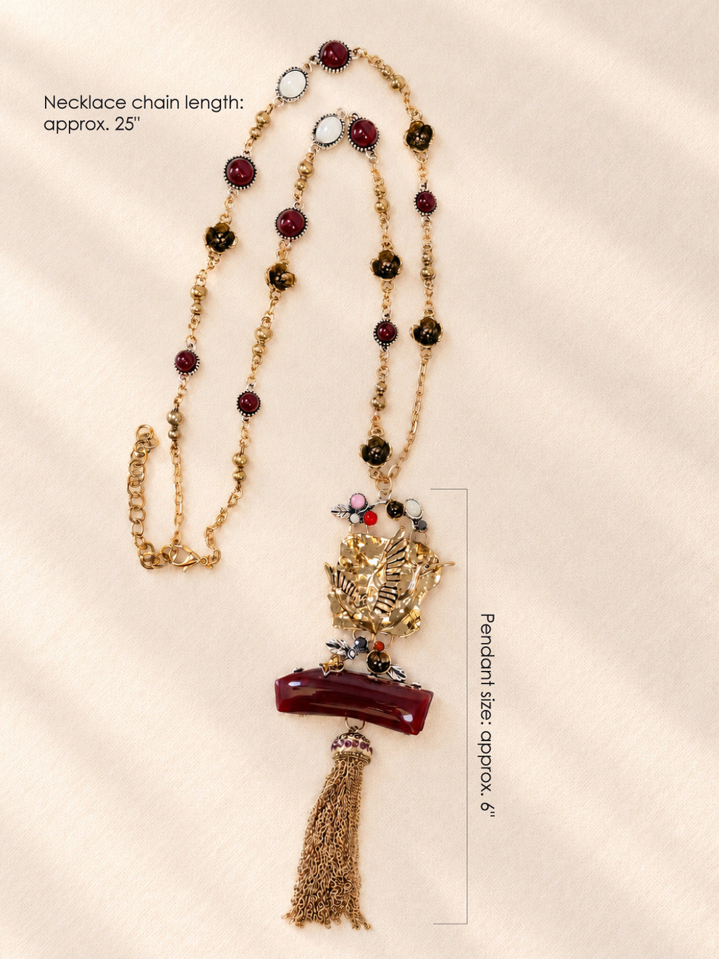 A long gold-toned beaded necklace with a detailed gold pendant featuring a bird motif, a dark red rectangular element, and a tassel, displayed on a cream satin fabric background with text overlays detailing measurements.