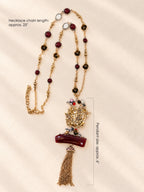 A long gold-toned beaded necklace with a detailed gold pendant featuring a bird motif, a dark red rectangular element, and a tassel, displayed on a cream satin fabric background with text overlays detailing measurements.