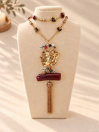A gold-tone layered necklace displayed on a cream jewelry bust. The necklace features a large central gold pendant with a stylized face, a horizontal dark red bar, a dangling gold tassel, and smaller red and black bead details on the chain.