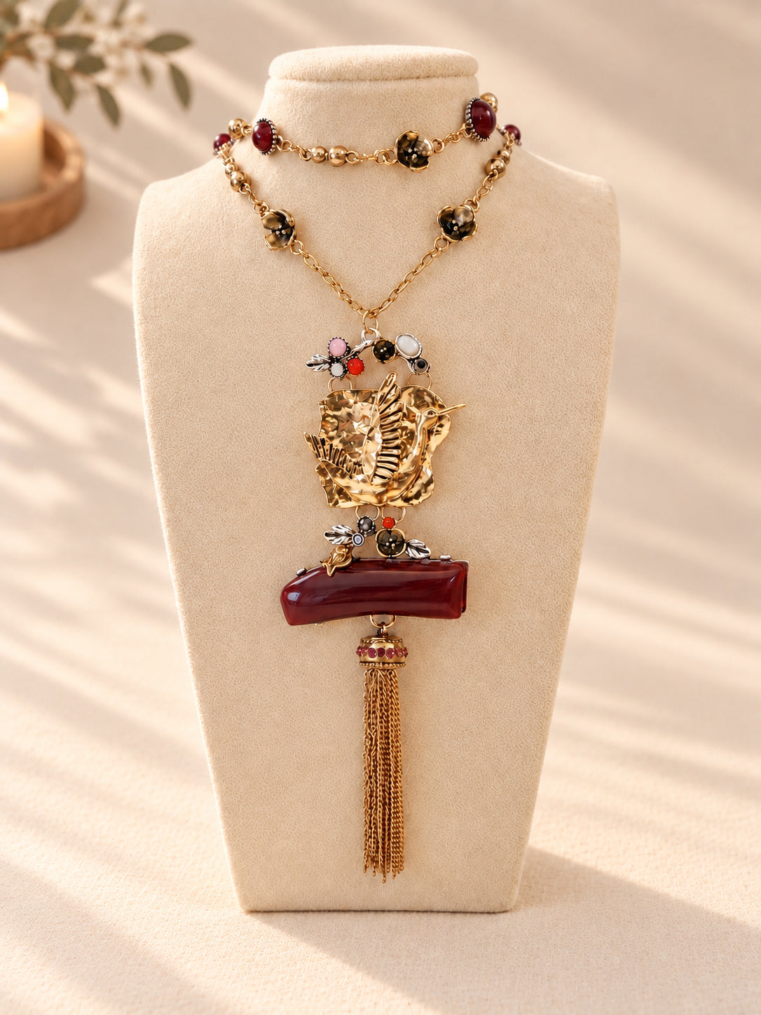 A gold-tone layered necklace displayed on a cream jewelry bust. The necklace features a large central gold pendant with a stylized face, a horizontal dark red bar, a dangling gold tassel, and smaller red and black bead details on the chain.