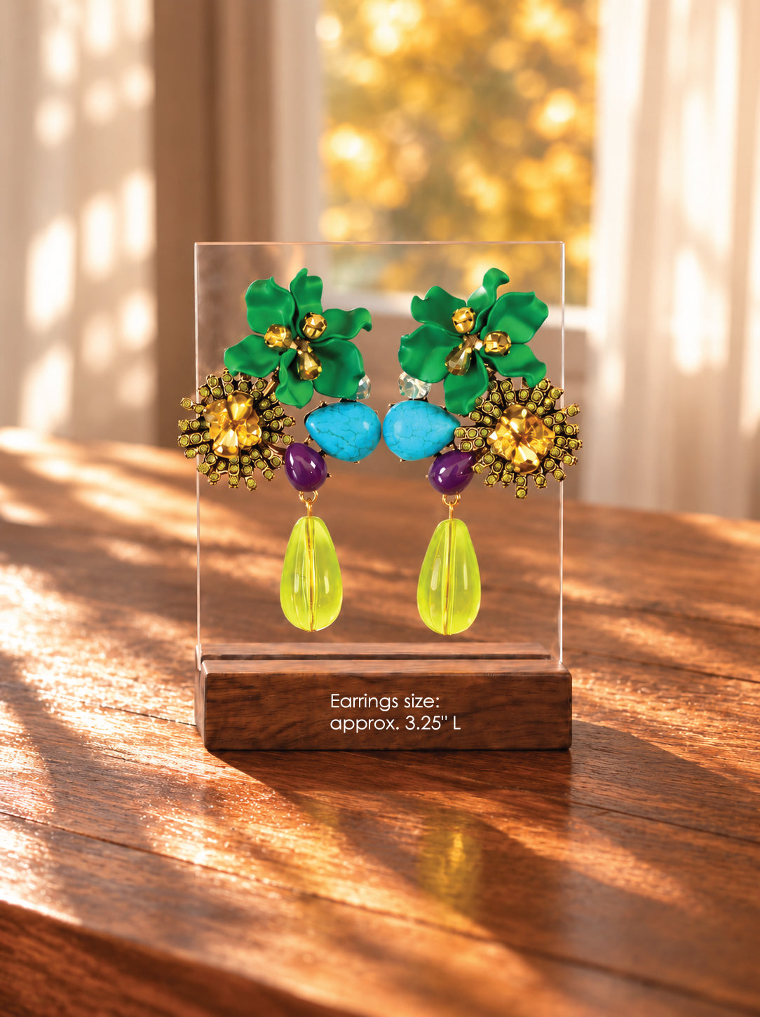A pair of colorful statement earrings displayed on a clear acrylic stand on a wooden table. The earrings feature green leaf-shaped studs with gold floral centers, light blue oval beads, purple spacer beads, and bright yellow-green teardrop drops.