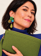 A woman wearing a textured olive green pebbled leather clutch bag and a matching green statement earring, styled with a blue ribbed turtleneck sweater against a light beige background.