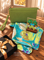 A curated gift set arranged on a wooden surface in warm sunlight, including an olive green crossbody pouch, a blue and green abstract print scarf, colorful floral earrings on a small wooden stand, and a black gift box with a gold ribbon. Text at the bottom reads the set includes crossbody bag, earrings, and scarf.