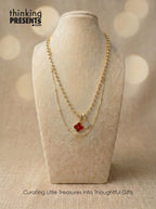 Layered gold-tone necklace with beaded chain and clover charm pendants in red and crystal detail, styled on a neutral display, elegant and feminine curated gift aesthetic