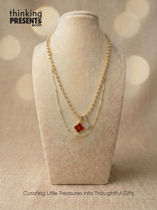 Layered gold-tone necklace with beaded chain and clover charm pendants in red and crystal detail, styled on a neutral display, elegant and feminine curated gift aesthetic