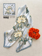Silky square scarf with lotus floral design in soft blue and ivory tones, paired with a vibrant orange scrunchie, styled as part of an elegant curated gift set by Thinking Presents™