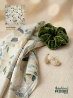 A styled flat-lay featuring a cream scarf with a muted green and gray botanical pattern, a dark green velvet scrunchie, pearl earrings, and a small inset showing the scarf design with text reading 'Scarf Design 25 in x 25 in'.