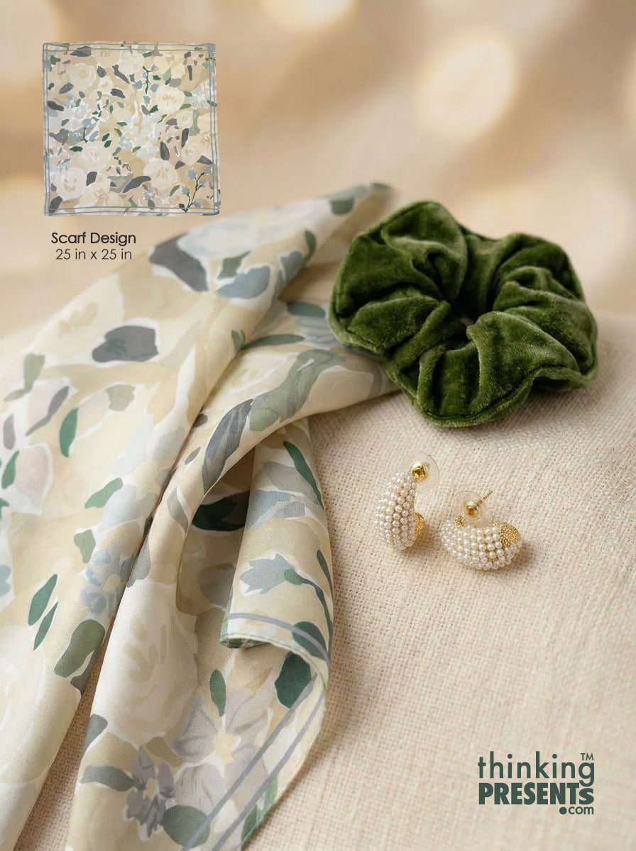 A styled flat-lay featuring a cream scarf with a muted green and gray botanical pattern, a dark green velvet scrunchie, pearl earrings, and a small inset showing the scarf design with text reading 'Scarf Design 25 in x 25 in'.