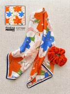 Floral scarf with orange and blue flowers on a beige background, accompanied by a matching scrunchie.