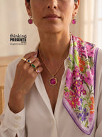 A close-up lifestyle shot of a model wearing a white blouse, a gold necklace with a magenta oval pendant, matching magenta earrings, and a colorful floral silk scarf. The text 'thinking PRESENTS.