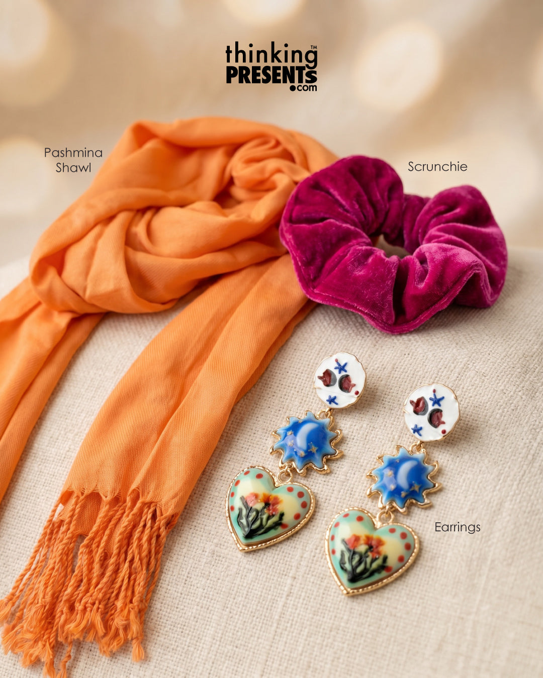 Orange shawl, pink scrunchie, and heart-shaped earrings on a beige surface with 'thinking PRESENTS' branding.
