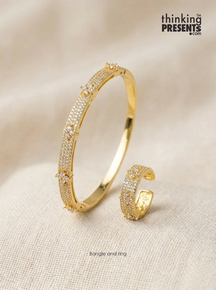 A gold-plated bangle bracelet and matching ring, both adorned with pavé-set clear stones, displayed on a textured beige fabric background. The brand logo 'thinking PRESENTS .