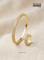 A gold-plated bangle bracelet and matching ring, both adorned with pavé-set clear stones, displayed on a textured beige fabric background. The brand logo 'thinking PRESENTS .