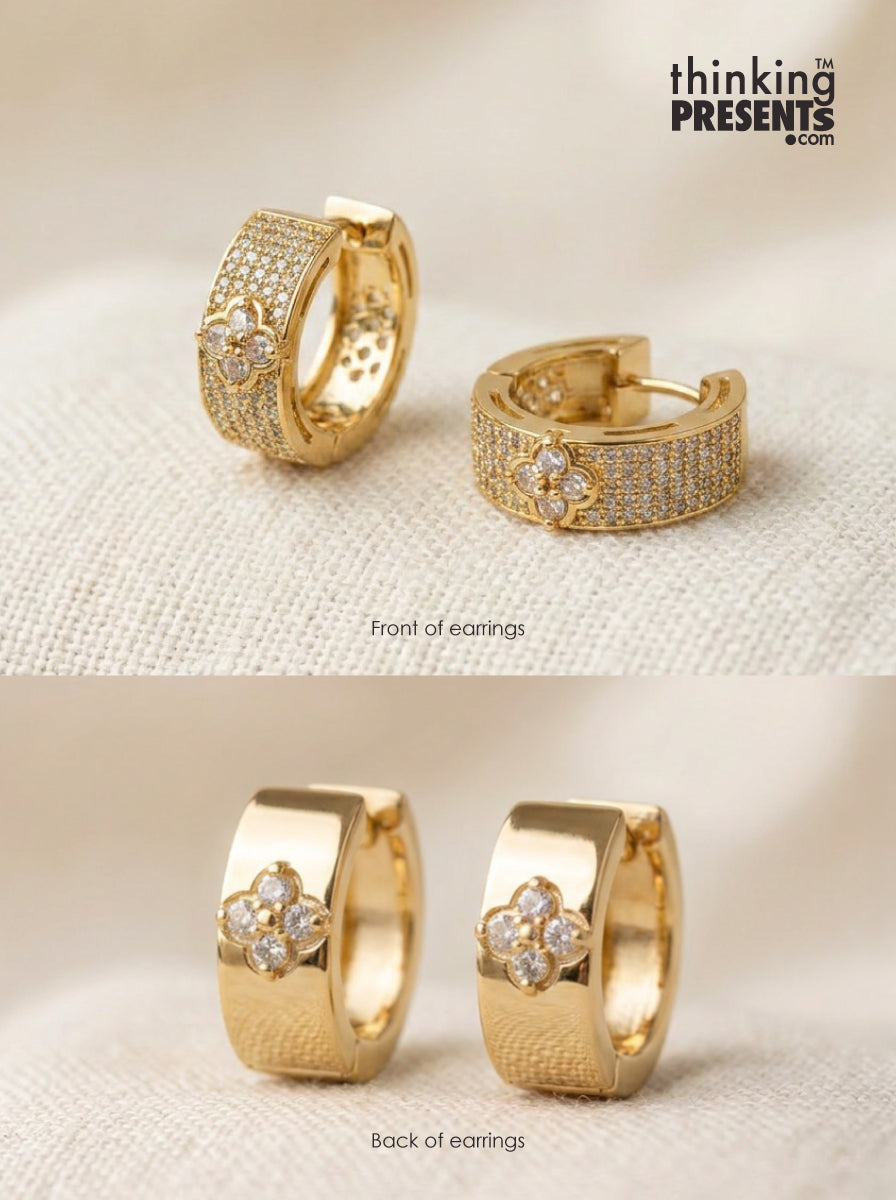 Gold huggie hoop earrings with pavé-set stones and a floral clover motif on the front, shown from both front and back views on a textured cream fabric background. The brand logo 'thinking PRESENTS .