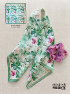 A floral silk scarf with green leaves and pink flowers on a white background, shown folded on a beige fabric surface next to a purple fabric accessory. A small inset at the top left shows the scarf design details with text reading 'Scarf Design 23 in x 23 in'.