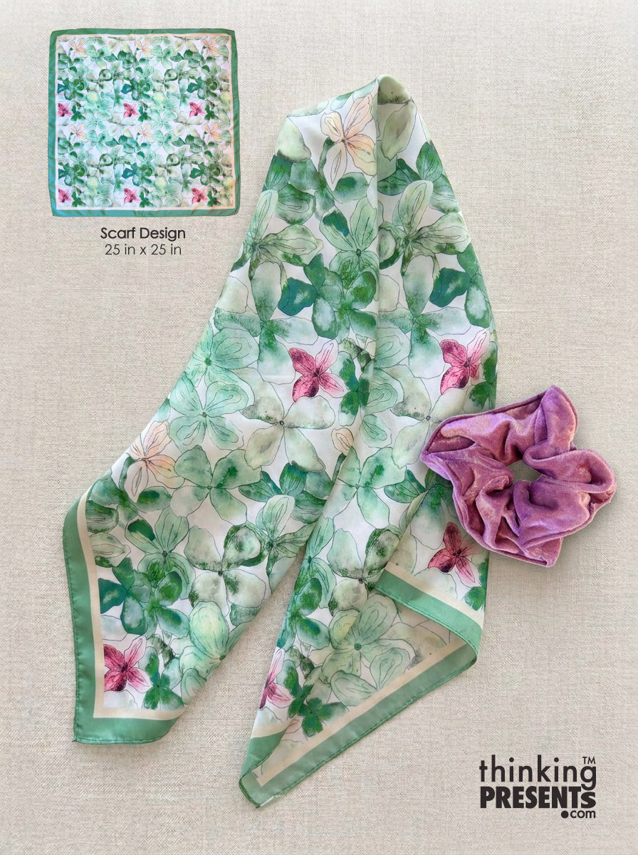 A floral silk scarf with green leaves and pink flowers on a white background, shown folded on a beige fabric surface next to a purple fabric accessory. A small inset at the top left shows the scarf design details with text reading 'Scarf Design 23 in x 23 in'.