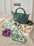 A styled gift set featuring a small green patterned handbag, a green and white floral scarf, a purple velvet scrunchie, and gold jewelry, arranged on a beige fabric surface with the text 'thinking PRESENTS.com' at the top.