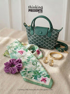 A styled gift set featuring a small green patterned handbag, a green and white floral scarf, a purple velvet scrunchie, and gold jewelry, arranged on a beige fabric surface with the text 'thinking PRESENTS.com' at the top.