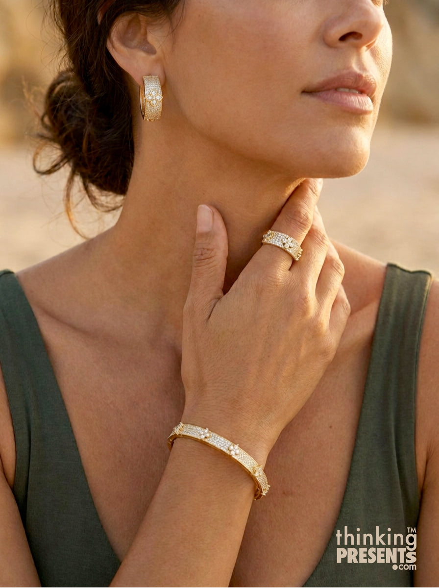 A close-up lifestyle shot of a woman wearing a gold cuff bracelet, matching gold hoop earring, and textured gold ring, set against a warm, blurred outdoor background.