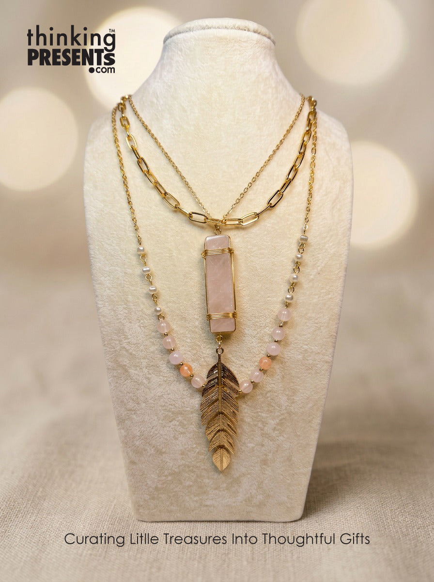Layered gold-tone necklace with blush pink stone bar pendant, soft beaded accents, and feather charm, styled on a neutral display, feminine and boho-inspired curated gift aesthetic