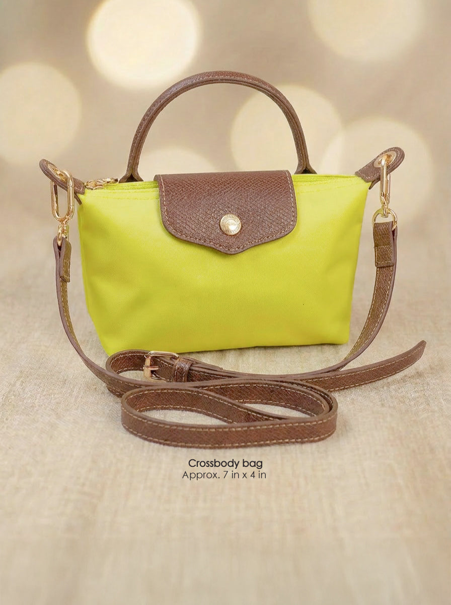 A small crossbody handbag with a bright lime green body and brown leather trim, featuring a top handle, a flap closure with a snap button, and a detachable shoulder strap, displayed on a beige textured surface with a warm bokeh background.