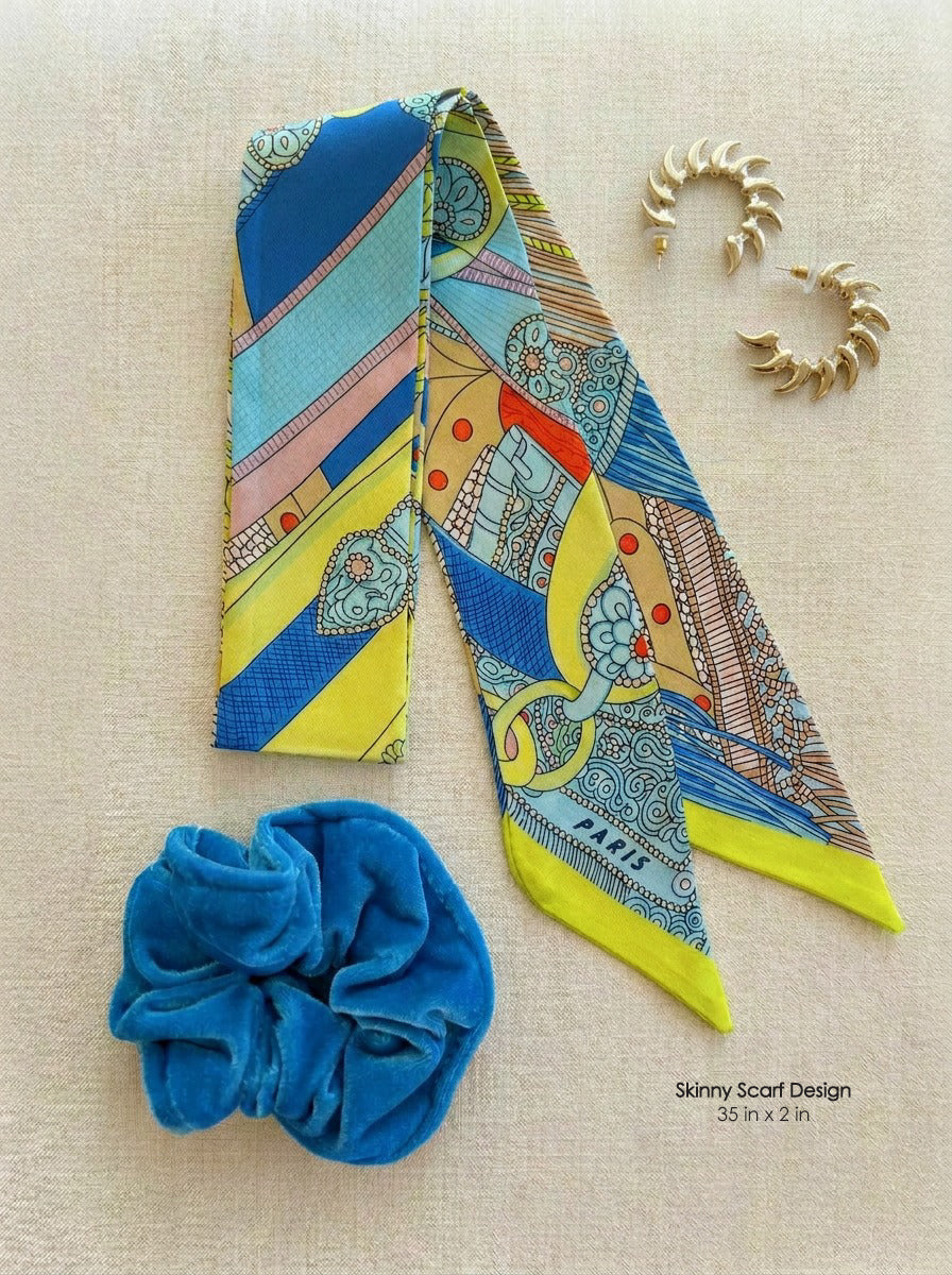 A colorful patterned scarf laid flat on a beige fabric background, shown with a bright blue velvet scrunchie and gold hoop earrings. Small text in the lower right reads 'Skinny Scarf Design 35 in x 2 in'.