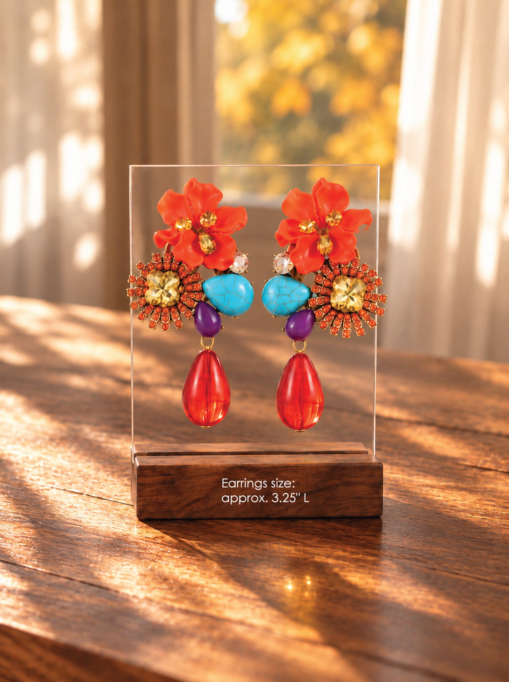 A pair of colorful statement earrings displayed on a clear acrylic stand with a wooden base on a wooden table. The earrings feature bright orange flowers, turquoise blue beads, purple beads, and red teardrop drops.